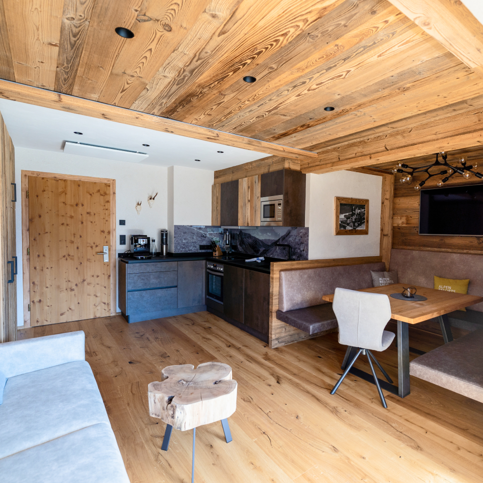 Rustic cabin interior with wooden ceiling and floor. Features a gray sofa, a carved wooden table with chairs, kitchen area with appliances, and a cozy dining nook with a cushioned bench. Natural light illuminates the modern yet homey space.