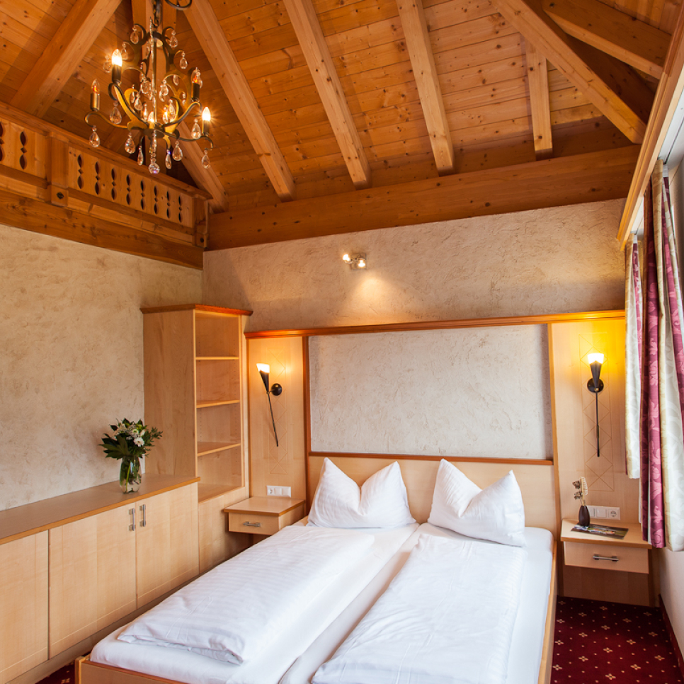 A cozy bedroom with a wooden ceiling and chandelier. It features a double bed with white bedding, wooden furniture, and red curtains. A vase of flowers is on the cabinet, adding a homely touch to the rustic decor.