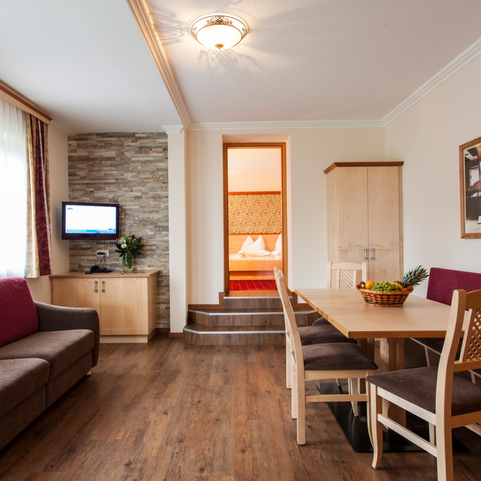 Cozy living room with wooden flooring, featuring a brown sofa, small TV on stone accent wall, and dining area with a table and chairs. A basket of fruit is on the table. Steps lead to a bedroom with a bed visible through a doorway.