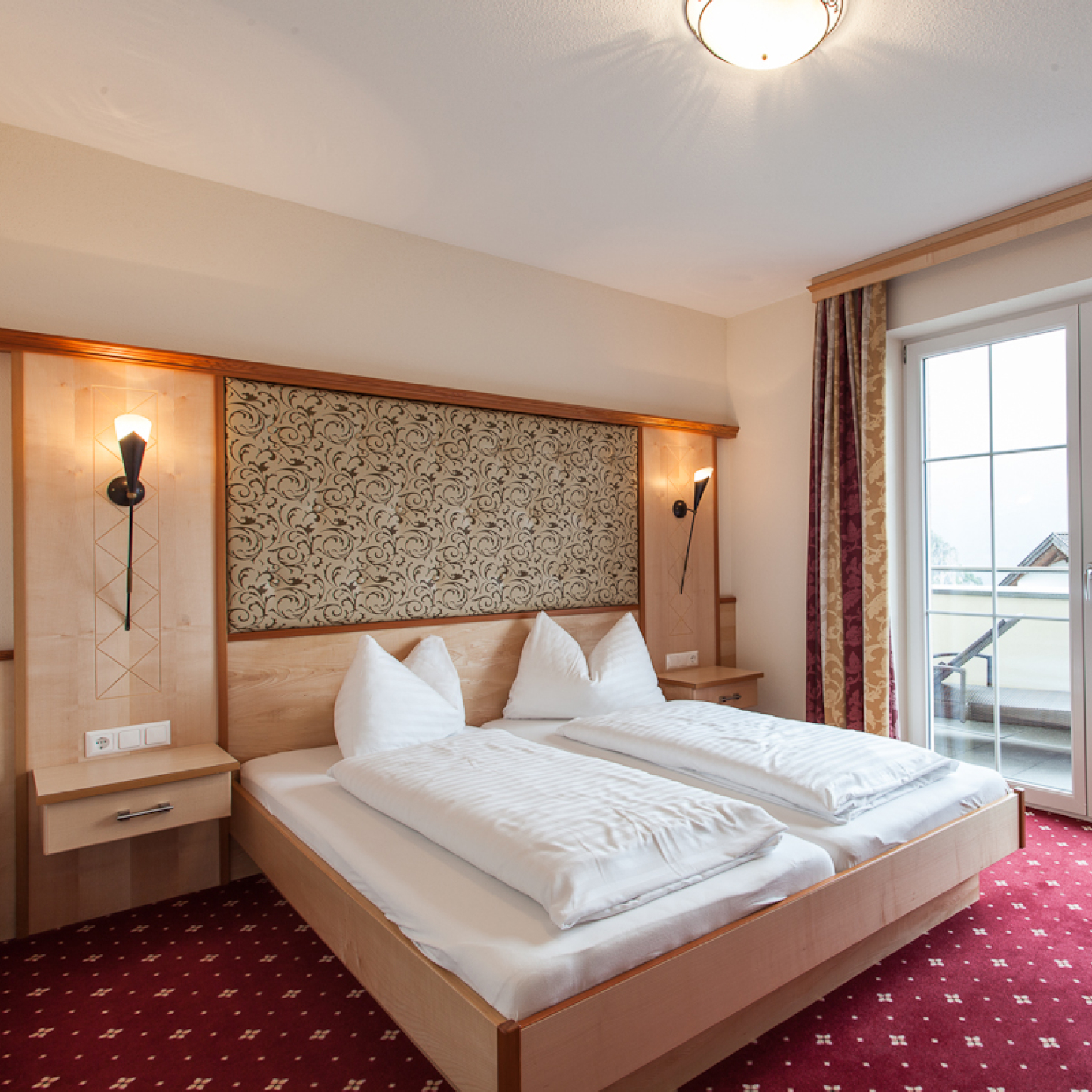 Bedroom with a double bed featuring two white pillows and sheets, wooden headboard with ornate design, wall-mounted lights, and a red carpet with a pattern. A glass door on the right opens to a balcony with outdoor scenery.
