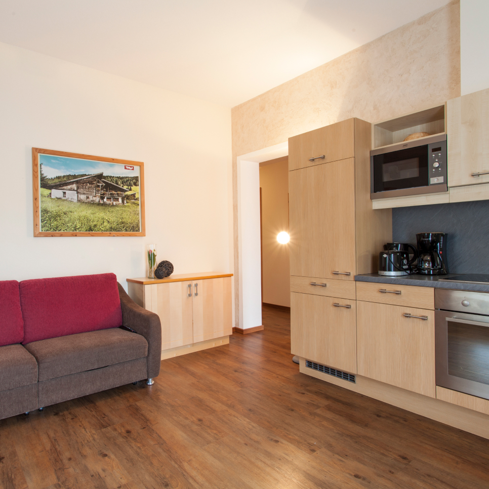 A modern living space with a brown and red sofa, wooden flooring, a kitchenette with light wood cabinets, oven, microwave, and coffee maker. A countryside photo hangs on a beige wall near a small wooden cabinet.