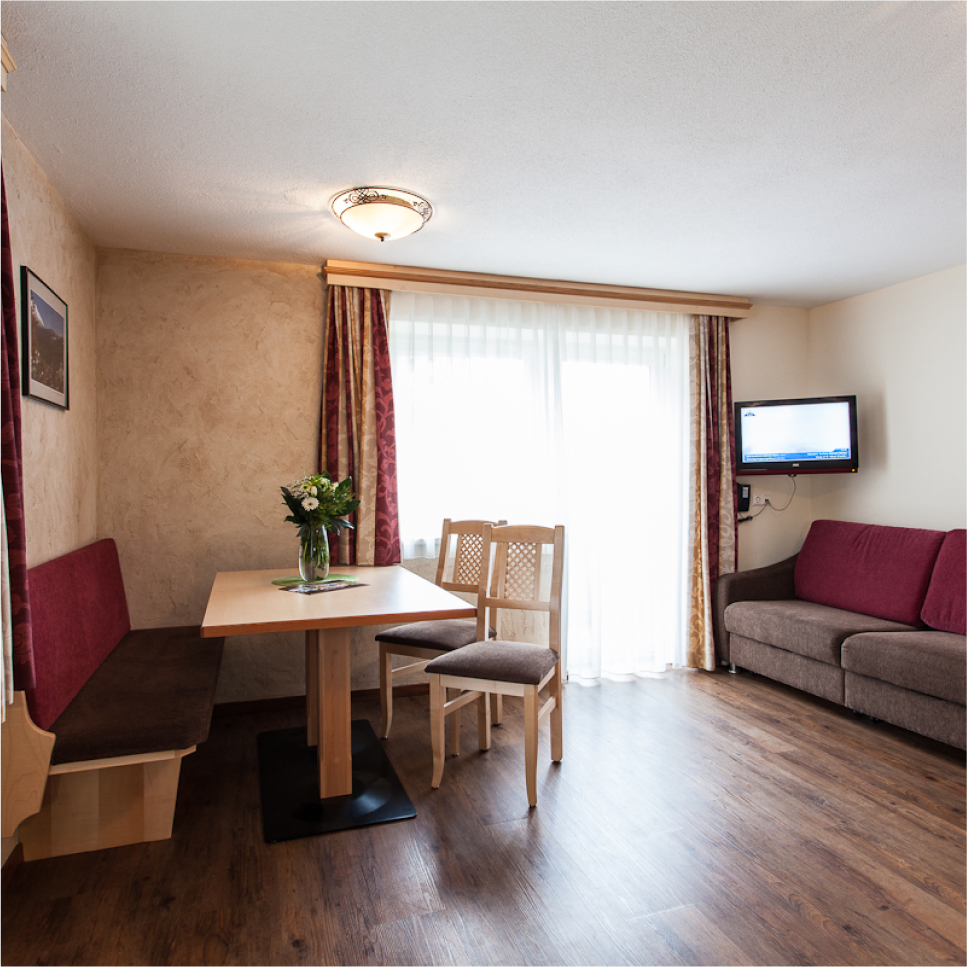 A cozy living and dining area with a wooden floor. It features a table with two chairs, a corner bench with red upholstery, a vase of flowers, a red sofa, a wall-mounted TV, and large curtained windows letting in natural light.