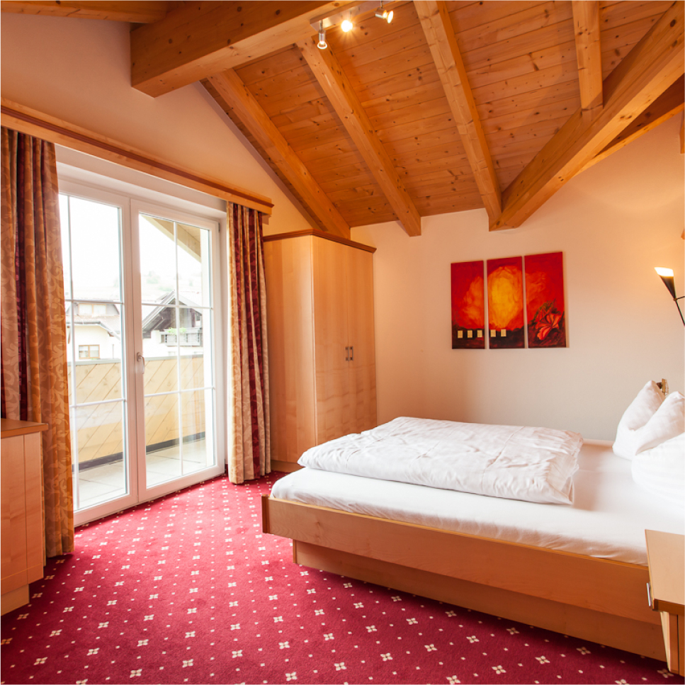 Cozy bedroom with a wooden ceiling, red carpet, and a double bed with white linens. There’s a balcony accessed through glass doors, and two red abstract paintings on the wall. Warm lighting creates an inviting atmosphere.