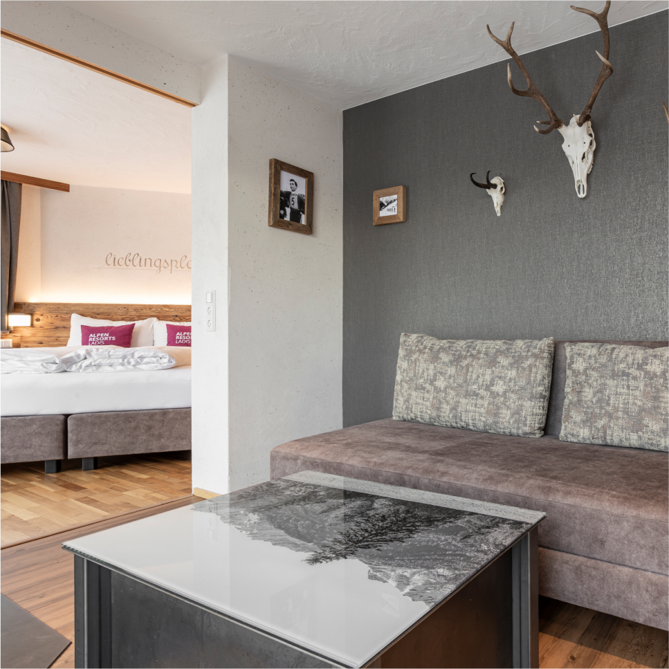 Modern hotel room with a large bed featuring white linens and pink pillows. A gray sofa sits by a glass-top table displaying a monochrome landscape. Antler decor and framed pictures adorn the walls, and wooden flooring adds warmth to the space.