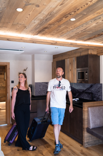 Eine Frau und ein Mann lächeln, als sie ein modernes Hotelzimmer im Chalet Ladis betreten. Sie tragen Koffer und sind offensichtlich gerade erst angekommen. Das Zimmer verfügt über Holzdecken, einen gemütlichen Küchenbereich und stimmungsvolles Licht, das sie herzlich willkommen heißt.