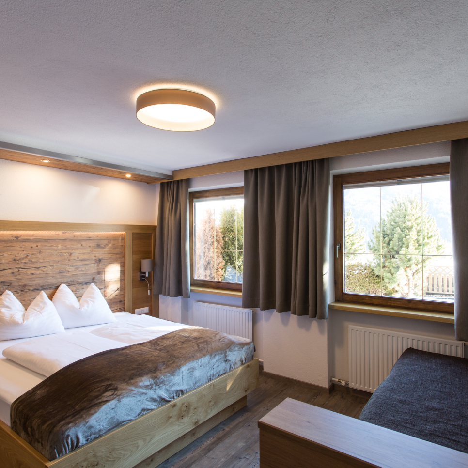 A cozy hotel room with a large bed, white linens, and wooden headboard. Two windows with gray curtains offer a view of trees and mountains. The room has wooden flooring, a modern ceiling light, and a small gray sofa.