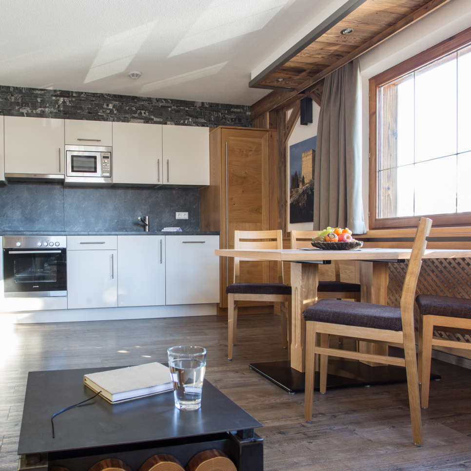Modern kitchen with white cabinets and stainless steel appliances, including an oven and microwave. A wooden dining table with chairs is near a large window, and a coffee table in the foreground holds a glass of water and a closed notebook.