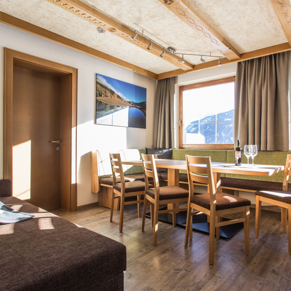A cozy dining area with a wooden table surrounded by chairs in a sunlit room. Theres a large window with a mountain view, a painting on the wall, and a bottle with two glasses on the table. The room has wooden floors and ceiling details.