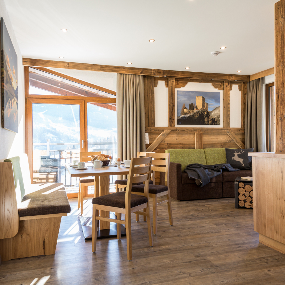 Cozy living area with wooden furniture and floors. A table with chairs is set for breakfast. Large windows offer a scenic mountain view. Art decorates light-colored walls. Firewood is neatly stacked beside a sofa with green cushions.