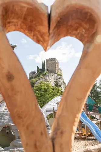 In der Ferne ist ein Steinschloss auf einem Hügel zu sehen, elegant eingerahmt von einer herzförmigen Holzkonstruktion. Darunter liegt ein Alpenresorts-Spielplatz mit Bäumen und einer Rutsche, teilweise sichtbar unter dem klaren blauen Himmel.