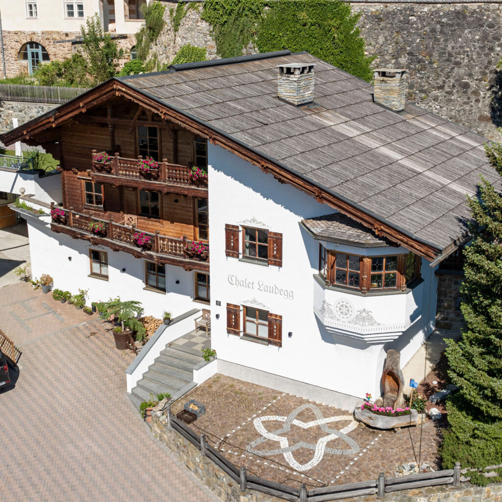 Chalet Laudegg in Ladis is a traditional retreat boasting wooden balconies and white walls. It features intricate carvings, vibrant flower boxes, and a circular stone patio with a star design, all enfolded by stone walls and lush greenery.