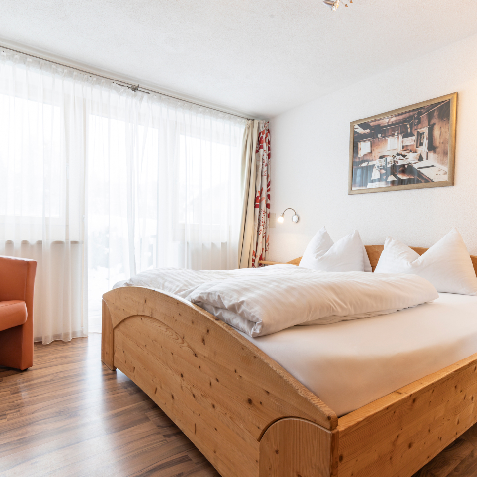 A cozy bedroom with a wooden double bed, white linens, and pillows. Large windows with sheer curtains allow natural light in. Theres a red armchair in the corner and a framed artwork on the white wall. The floor is wooden.