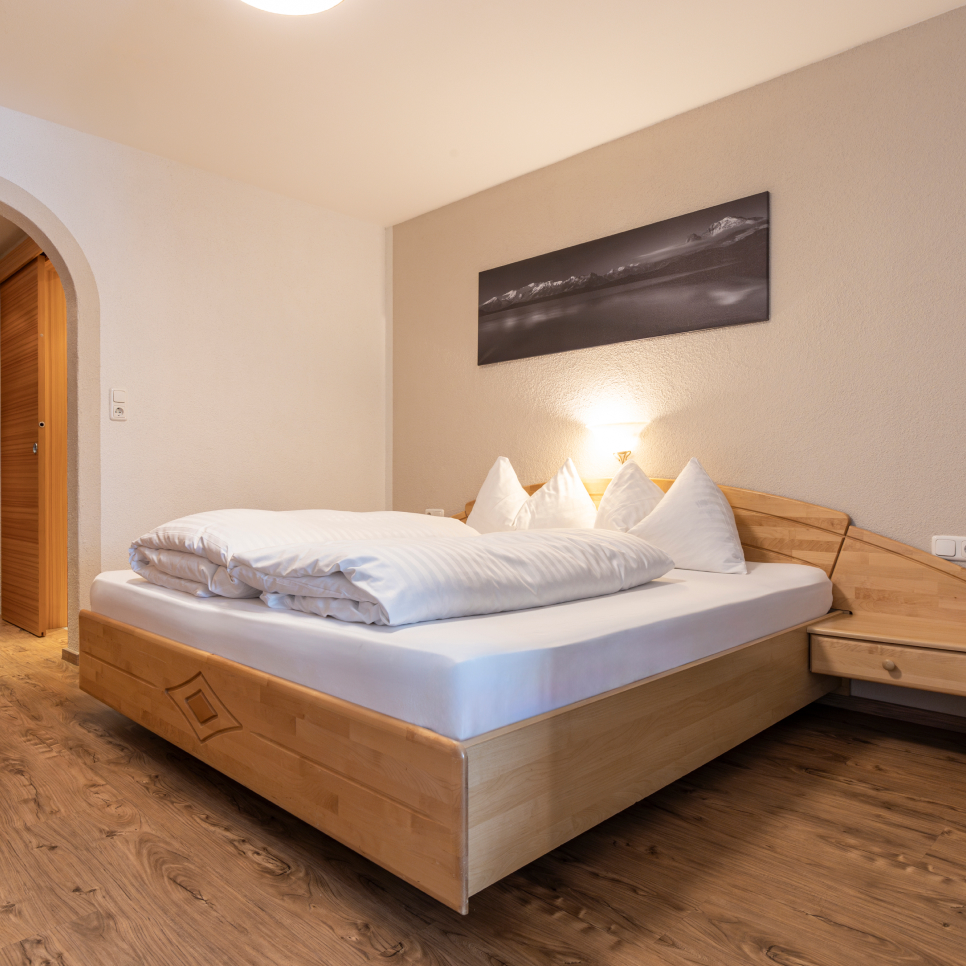 A cozy bedroom with a wooden bed featuring white bedding and pillows. A black and white landscape photo hangs above the bed. A wooden nightstand is on the right, and the room has wooden flooring and an arched doorway.