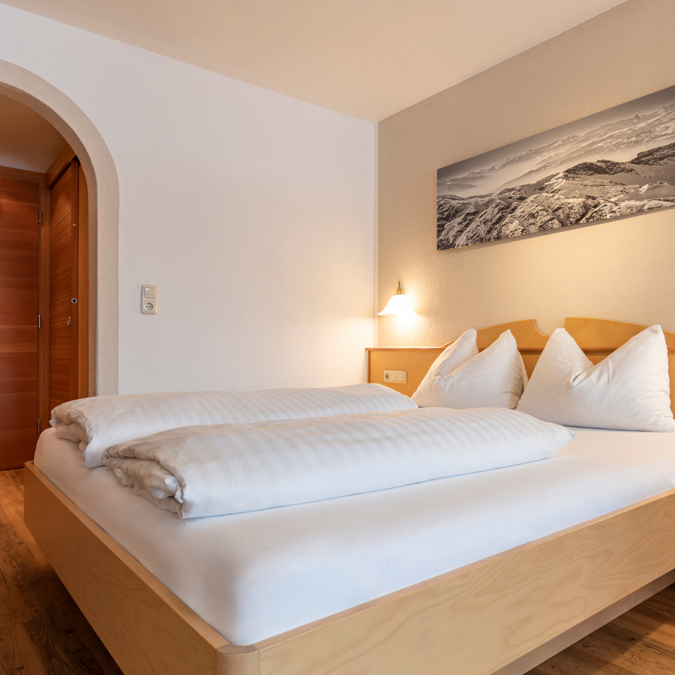 A cozy bedroom with a wooden double bed, white bedding, and pillows. A soft yellow wall lamp is lit above the bed. A black-and-white mountain landscape photo hangs on a light-colored wall. A wooden door and archway are visible.