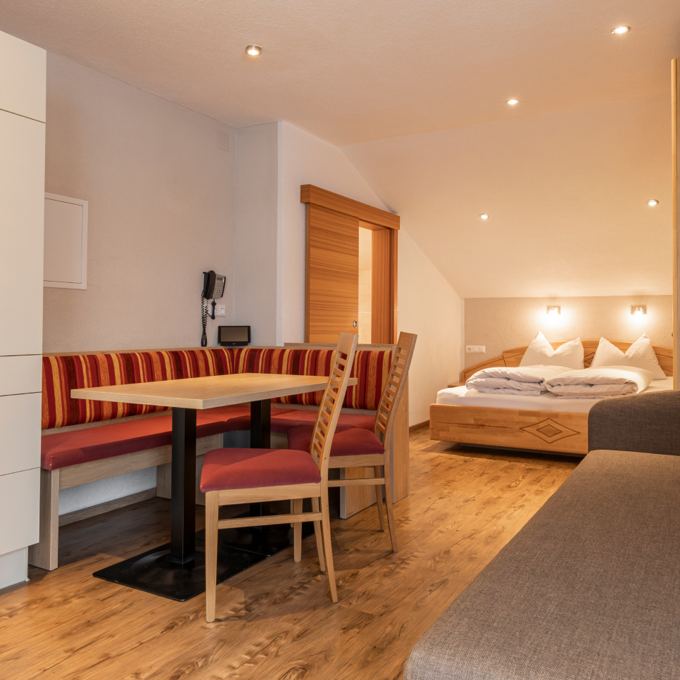 A cozy studio apartment with wooden flooring, featuring a double bed, a small dining area with a wooden table and red upholstered chairs, and a built-in bench. Soft lighting and a sliding wooden door complete the warm ambiance.