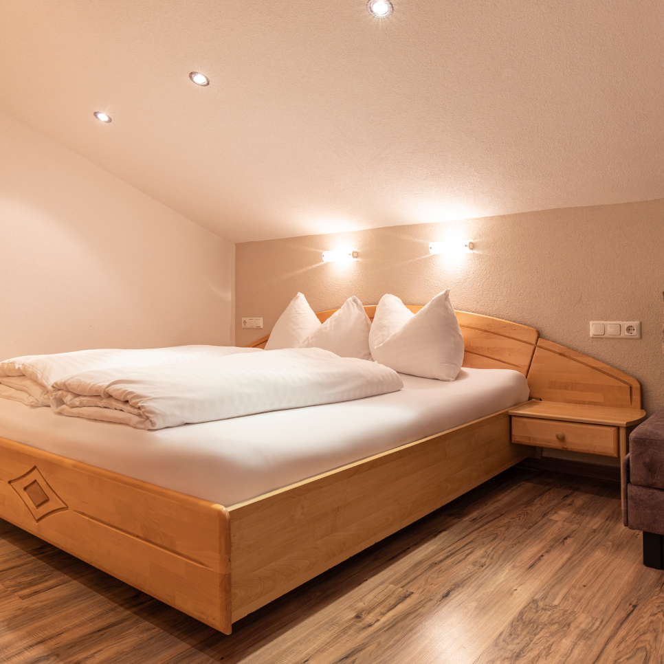 Cozy bedroom with sloped ceiling, featuring a wooden bed with white bedding, two pillows, and a folded blanket. Warm lighting from recessed ceiling lights and two wall lamps illuminates the wooden floor and small bedside table.