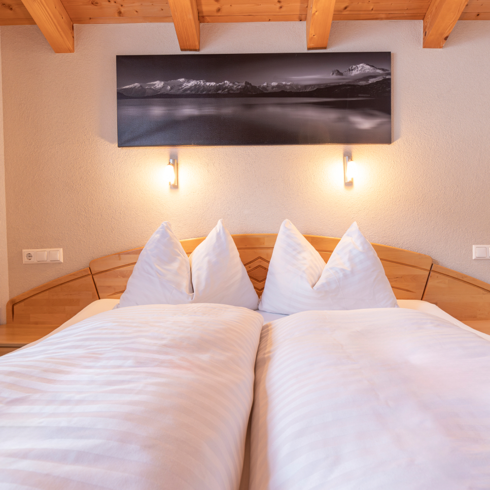 A neatly made bed with white pillows and a duvet is positioned under a photograph of mountains above a lake. Two wall-mounted lights illuminate the scene, and wooden beams are visible on the ceiling, creating a cozy atmosphere.