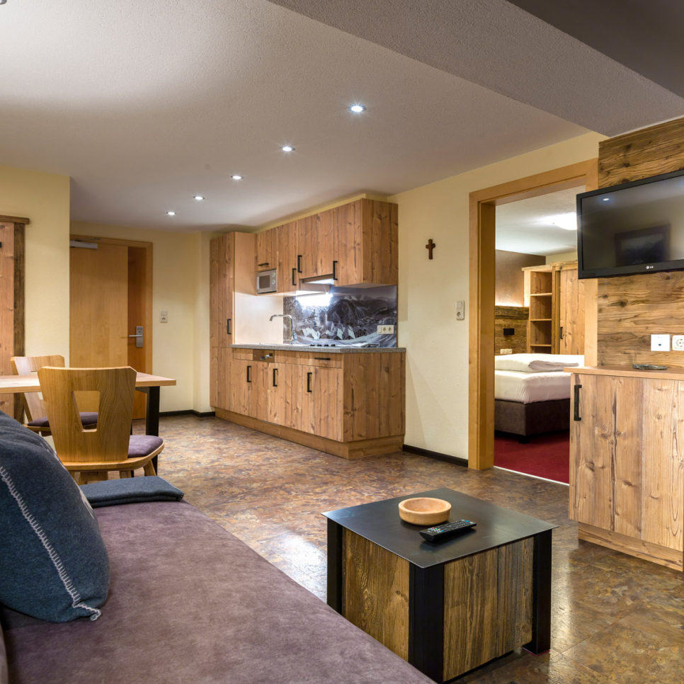 A cozy living room with a sofa, a small black coffee table, and a flat-screen TV on the wall. Nearby is a kitchenette with wooden cabinets. The room has a warm, rustic décor, and a wooden dining table with chairs. A bedroom is visible in the background.