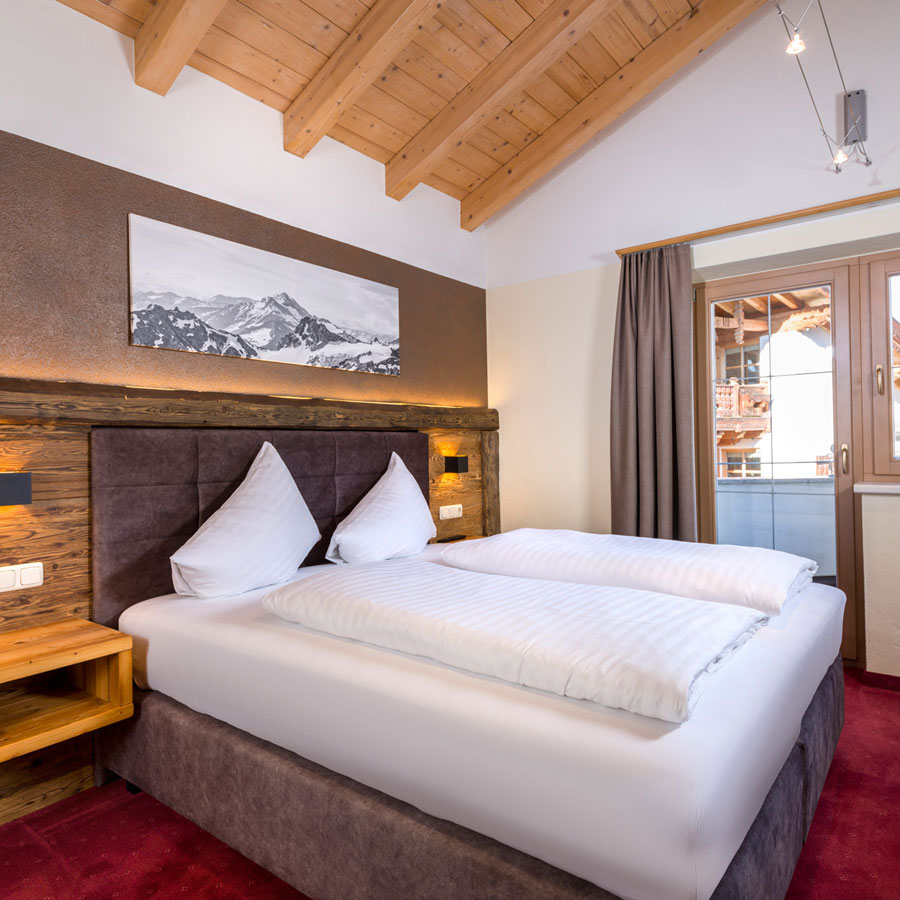 A cozy bedroom with a wooden beamed ceiling, featuring a large bed with white linens and two pillows. The headboard is made of dark wood. A landscape picture hangs above, and a large window with a curtain is on the right. Red carpet covers the floor.