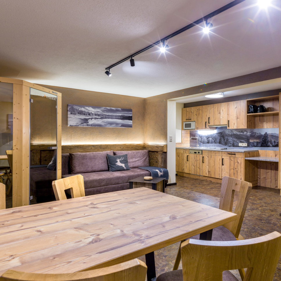 Cozy dining and living area featuring a wooden table with chairs, a brown sofa adorned with deer-themed cushions, and a wooden kitchenette. The room is warmly lit with track lighting and decorated with nature-themed wall art.