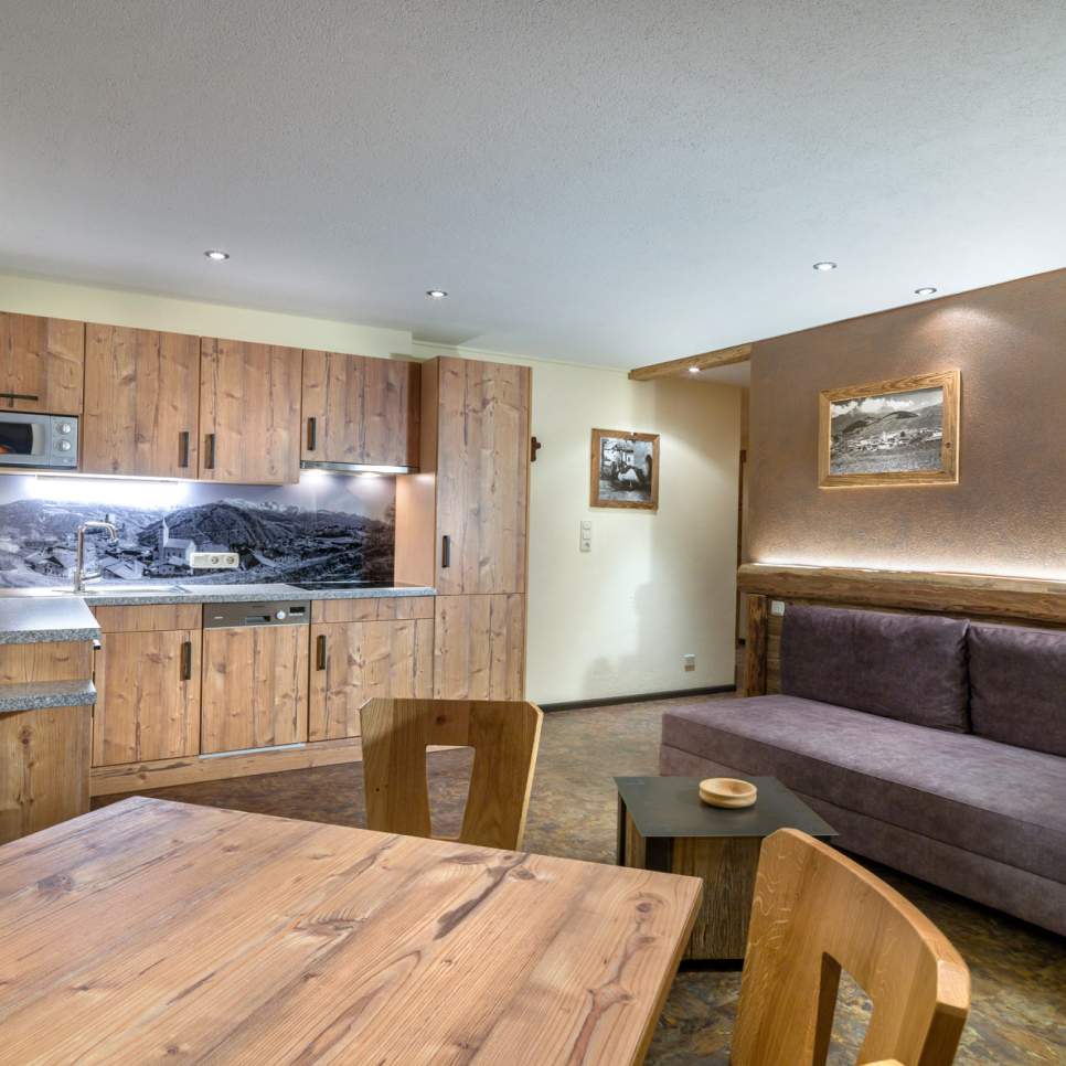A cozy living area features a kitchenette with wooden cabinets, a black and white backsplash, and a microwave. Theres a gray sofa with framed pictures above it, a wooden dining table with chairs, and a small coffee table. Neutral walls complete the space.