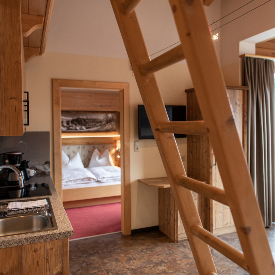 Dieses gemütliche Apartment aus Holz, ideal für 4-5 Personen in Ladis, verfügt über eine Leiter, die zu einem charmanten Dachboden führt. Darunter befindet sich die Küche mit Spüle und Kaffeemaschine. Hinter einer offenen Tür liegt ein Schlafzimmer mit einem ordentlich gemachten Bett und Kissen, während Vorhänge das hohe Fenster wunderschön einrahmen.