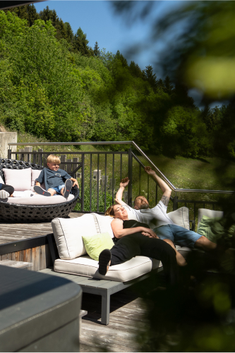 A family relaxes on a sunlit outdoor deck surrounded by greenery. A woman reclines on a couch with a man lying beside her, both looking content. A child sits nearby in a round chair, smiling and enjoying the sunny day.