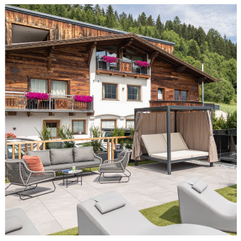 Outdoor terrace with modern seating including sofas and loungers. A cabana with curtains is on the side. The backdrop features a rustic wooden chalet adorned with pink flowers on balconies. Lush greenery and trees are visible in the background.