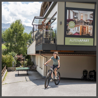 Eine Frau fährt mit dem Fahrrad eine Einfahrt in der Nähe eines Gebäudes mit einem großen Werbeplakat für „Alpen Apart Alexander“ hinunter. Im Hintergrund sind Grünflächen und Berge zu sehen, und neben der Einfahrt sind ein Tisch und zwei Kinderwagen zu sehen.