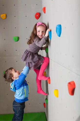 Ein junges Mädchen in einem grauen Kleid und rosa Leggings klettert eine Indoor-Kletterwand mit bunten Griffen hoch. Ein Junge in einer blauen Jacke hilft, indem er ihr Bein hält. Beide Kinder lächeln und scheinen Spaß zu haben.