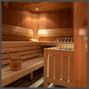 Eine warme, einladende Sauna mit Holzbänken und -wänden. Auf einer unteren Bank stehen Eimer und Schöpfkelle, während in der Mitte ein Metallofen mit heißen Steinen steht. Sanftes Licht sorgt für eine entspannende Atmosphäre.