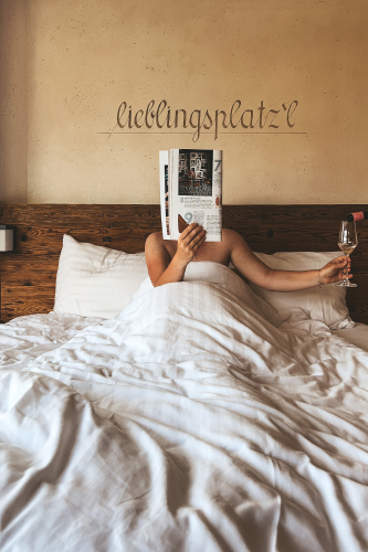 Person sitting in bed under white sheets, holding a magazine in front of their face and a wine glass in the other hand. The headboard has the word lieblingsplatzl written on it. The setting appears cozy and relaxed.