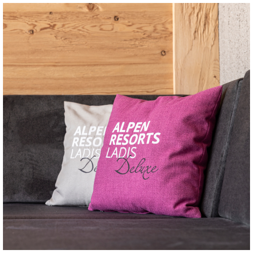 Two throw pillows are placed on a dark sofa against a wooden wall. The left pillow is light gray with ALPEN RESORTS LADIS Deluxe text, and the right pillow is dark pink with a similar text design.