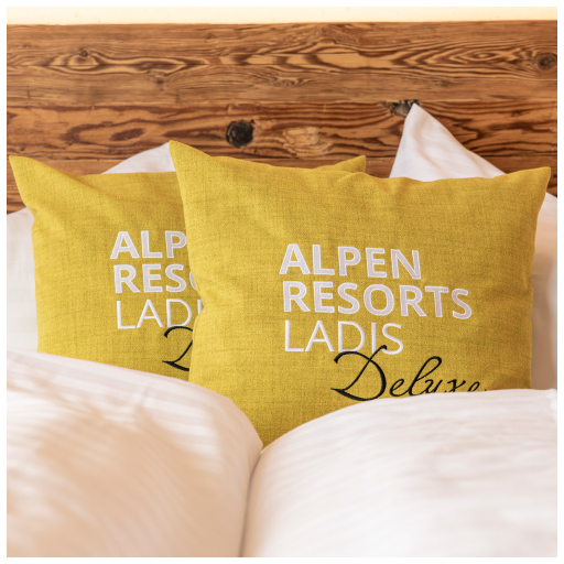 Two yellow throw pillows with Alpen Resorts Ladis Deluxe embroidered on them, placed on a made bed with white linens. A rustic wooden headboard is visible in the background.