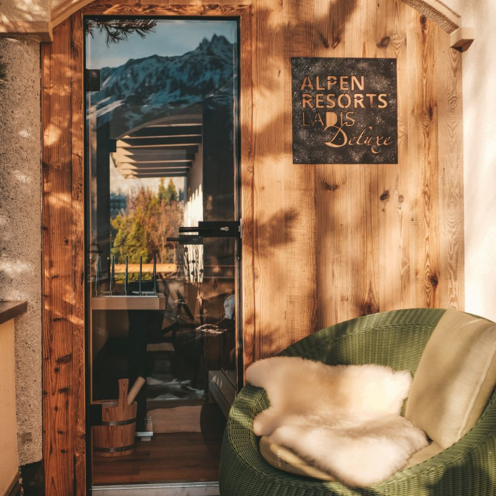 A cozy outdoor seating area features a green cushioned chair with a fur throw, next to a wooden door with a window. A sign above reads Alpen Resorts LADIS Deluxe. Sunlight and shadows create a warm, inviting atmosphere. Snow-capped mountains are visible.
