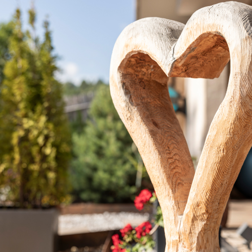 Inmitten der ruhigen Umgebung des Garten Spa mit Whirlpool im Laudegghof steht im Freien eine herzförmige Holzskulptur, umrahmt von einer verschwommenen Kulisse aus üppig grünen Bäumen und leuchtend roten Blumen an einem sonnigen Tag.