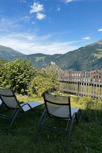 Two lounge chairs on a grassy lawn facing a scenic view of mountains. A wooden fence and bushes are in the background, with a distant castle visible on a hill under a clear, blue sky with a few clouds.