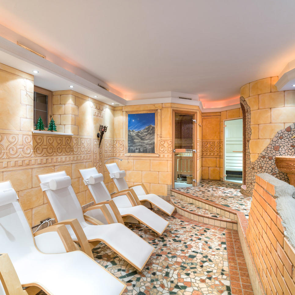 A cozy spa room with four white lounge chairs lined up side by side. The flooring features a mosaic tile design, and the walls are adorned with warm-toned tiles. A painting and two small decorative trees are displayed on the wall.