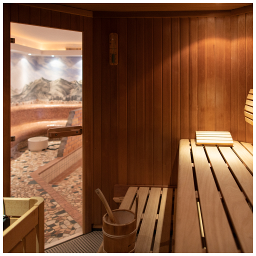A wooden sauna interior with seating benches and a bucket in the foreground. The door is open, revealing a tiled room with mountain-themed wall art in the background. Soft lighting creates a warm, inviting atmosphere.
