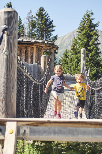 Two children, a boy and a girl, happily run across a wooden suspension bridge. The bridge is flanked by trees and a wooden structure, with a mountain in the background under a clear sky. They are smiling and wearing casual summer clothes.
