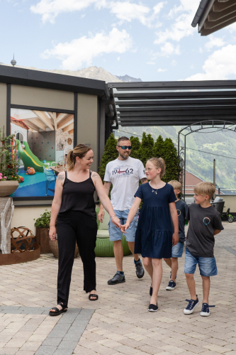 A family of four walks outdoors on a sunny day, with mountains in the background. The mother and daughter hold hands, while the father and son walk beside them. The setting includes potted plants and a building with a mural.