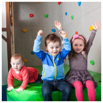 Three children are sitting on a green padded platform in front of an indoor climbing wall with colorful holds. The two children on the right, wearing a blue jacket and a gray dress, have their hands raised. The child on the left, in red, is looking forward.