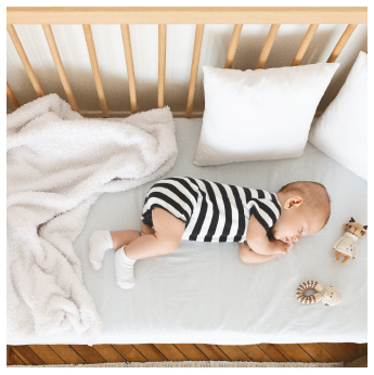 A baby is sleeping on a white bed in a wooden crib, wearing a black and white striped onesie and white socks. A soft blanket and two plush toys, an animal and a doll, are near the baby.