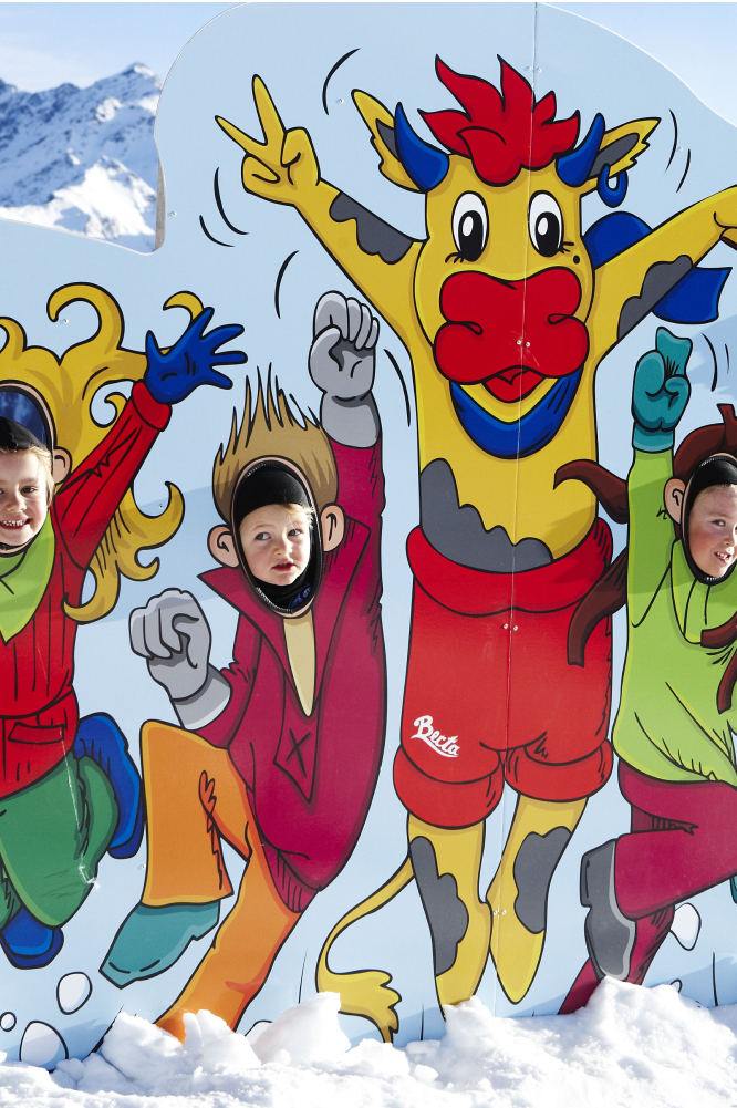 Children playfully pose behind a colorful cutout featuring cartoon characters, including a cow in a cheerful dancing pose. Snowy mountains are visible in the background, suggesting a winter setting.