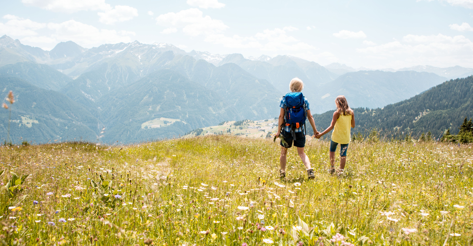 Two people are walking hand in hand through a wildflower meadow on a sunny day, with a panoramic view of mountains in the background. One person carries a backpack, and both are wearing summer clothing. The scene is peaceful and scenic.