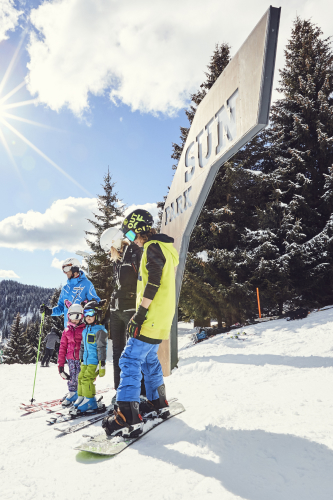 A group of people, including four children and one adult, stand in the snow with skis and snowboards. They are wearing helmets and winter gear. Behind them is a large metal sign reading SUN PARK, and pine trees are visible under a sunny sky.