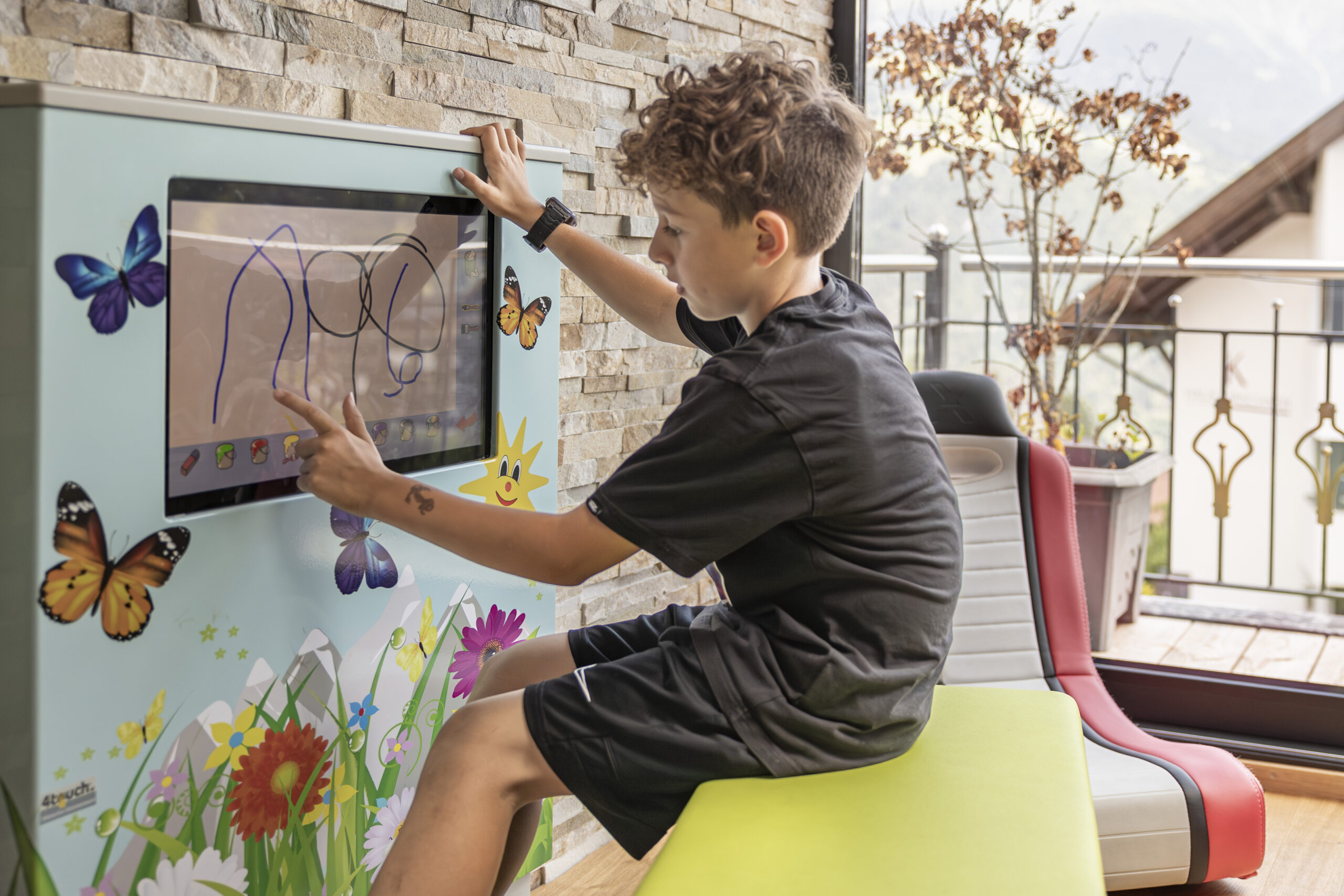 Junge malt auf einem Touchscreen in Mimis Kinderspielwelt im Alpenapart in Ladis, Zimmer mit Schmetterlingsmotiv.
