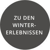 Ein dunkelgrauer Kreis mit weißem Text auf Deutsch lautet „ZU DEN WINTER-ERLEBNISSEN“ und lädt Sie ein, Alpenresorts zu erkunden.