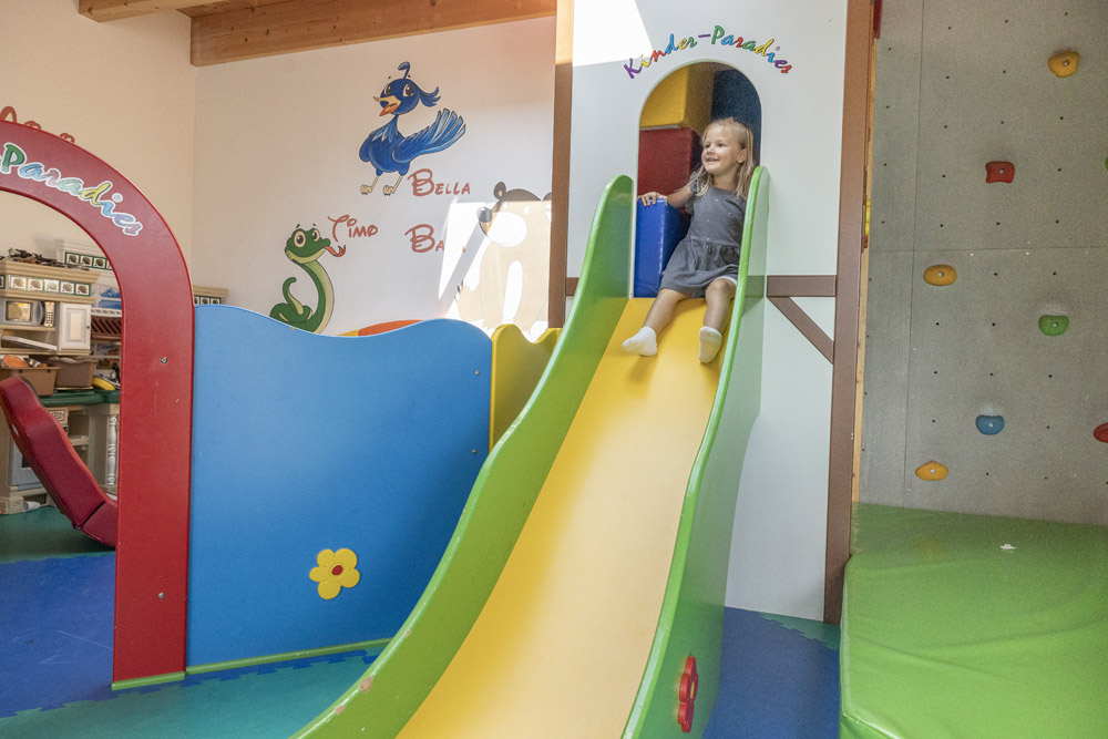Ein kleines Mädchen in einem grauen Kleid genießt die gelbe Rutsche in Mimis Kinderspielwelt im Alpenapart.