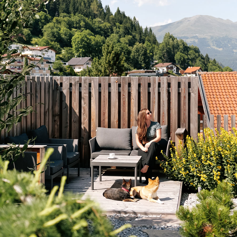 A woman sits on a patio sofa in a garden, looking away, with two dogs lying at her feet. The area is surrounded by wooden fencing and lush greenery. In the background, there are houses on a hillside and a mountain under a clear blue sky.